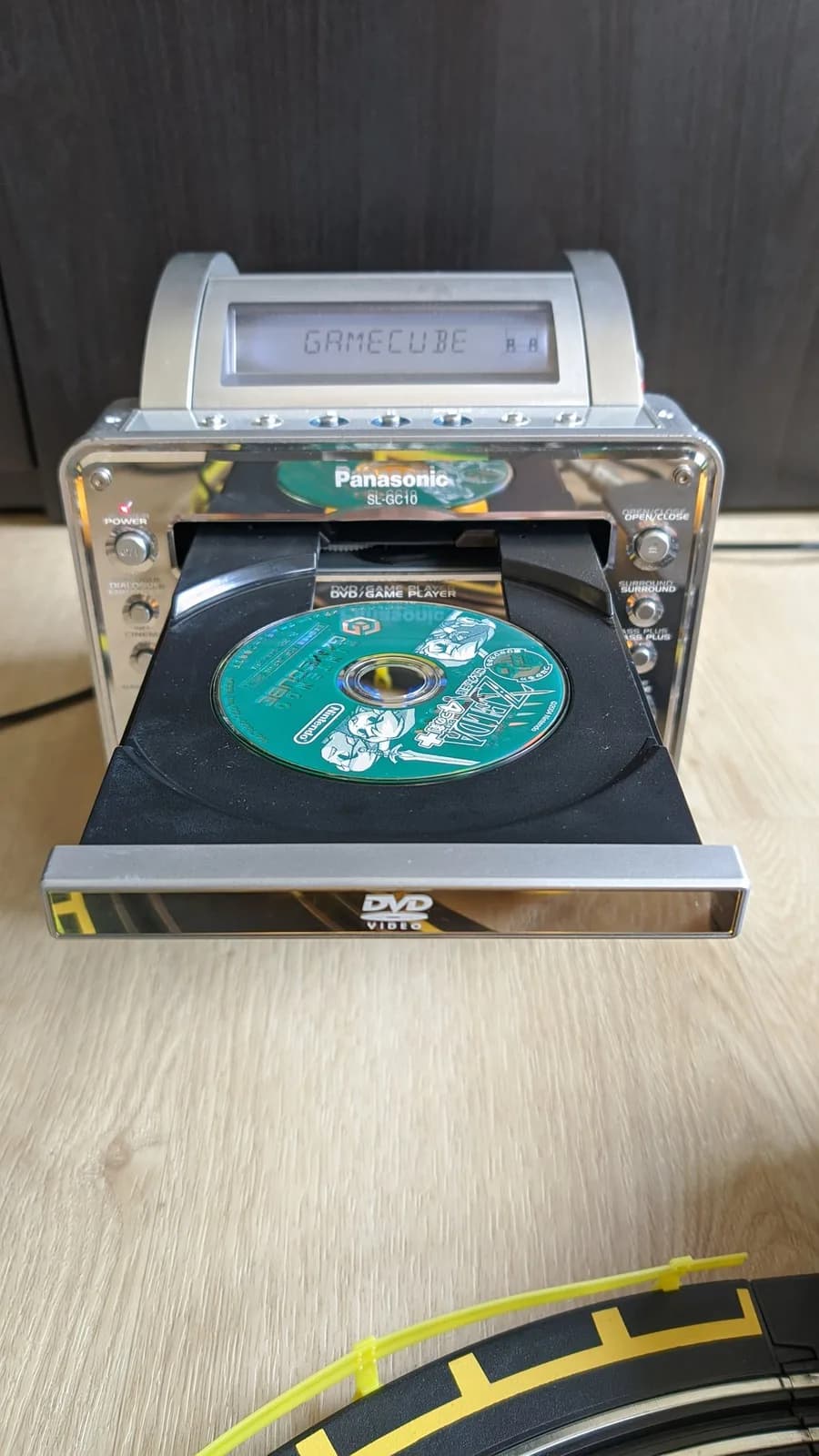 Panasonic Q with open DVD drive tray showing green circuit board and disc, LCD display reading "GAMECUBE", transparent housing exposing internal components.