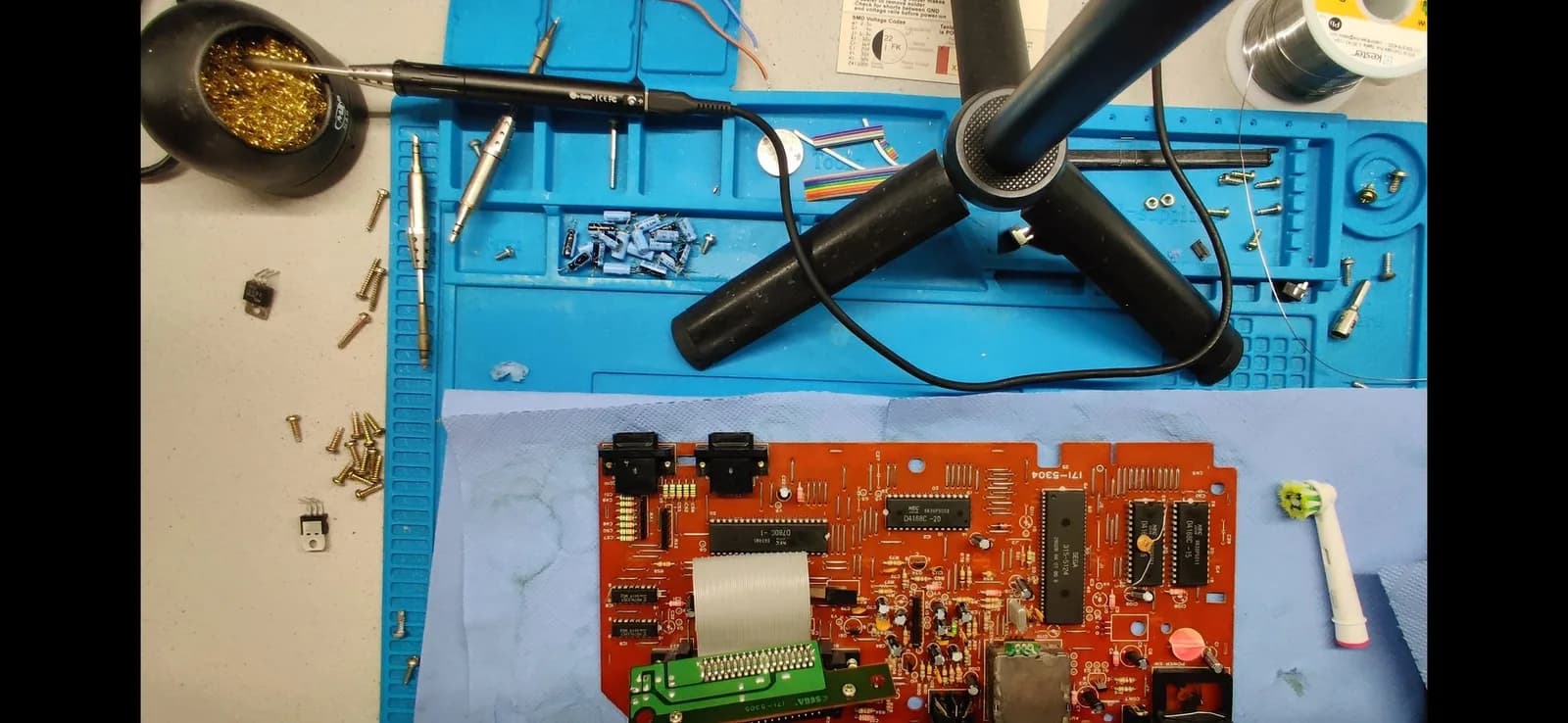 Workbench view of the red Sega Mark III motherboard (silkscreen 171-5904) on a blue silicone mat, recapped with the new capacitors fitted. Off to the left on the bench: the old removed capacitors, the new 7805 voltage regulator and the shell's screws, with a soldering iron, desoldering tool and solder to hand.