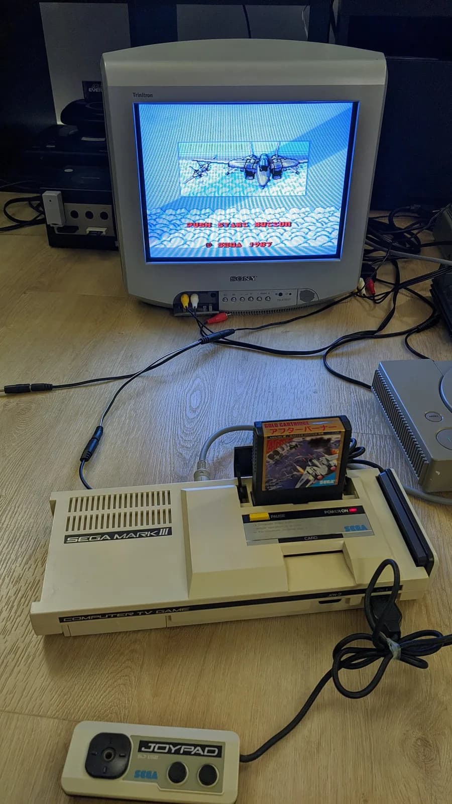 Sega Mark III on a wooden floor connected to a Sony Trinitron CRT TV showing the After Burner title screen (jet over clouds, PUSH START BUTTON, SEGA 1987), the gold After Burner Sega card in the top slot and a Sega Joypad controller in front.