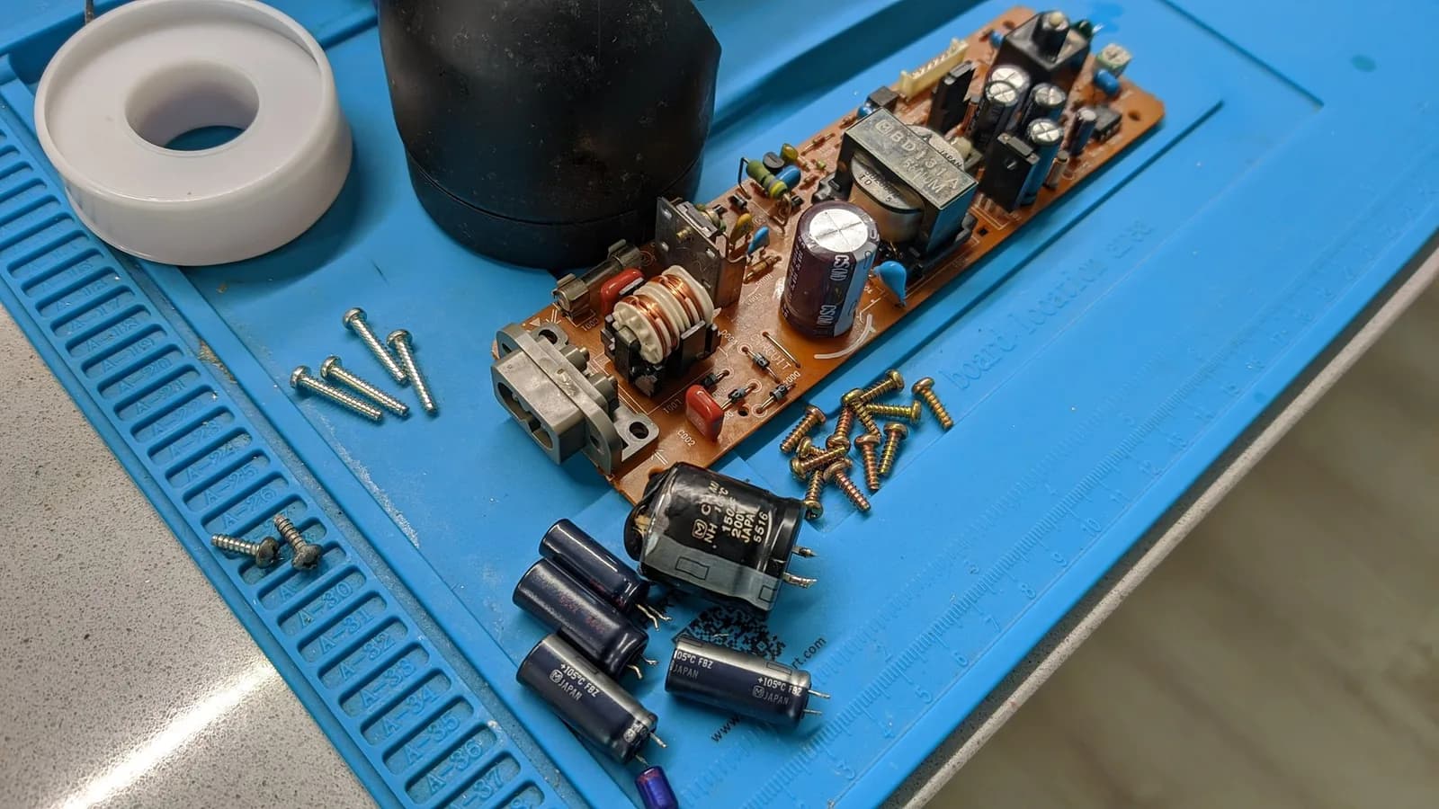 PS1 PSU board, removed from its shell and freshly recapped, on a blue silicone mat with the transformer and coil still mounted. Around it on the mat are the old capacitors removed during the recap, including the main filter cap with its top ruptured from venting, plus scattered screws.