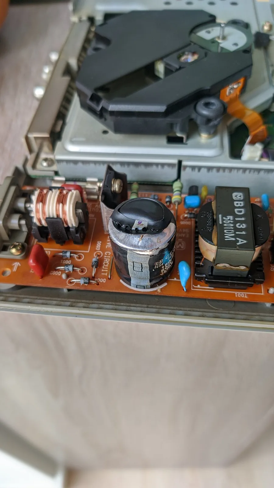 Close-up of the PS1 PSU board with the big main filter cap blown and vented, its foil wrap split open. The filter cap was the only damaged component; the transformer beside it is fine and the orange PCB is undamaged. The CD drive optical pickup sits above the board.
