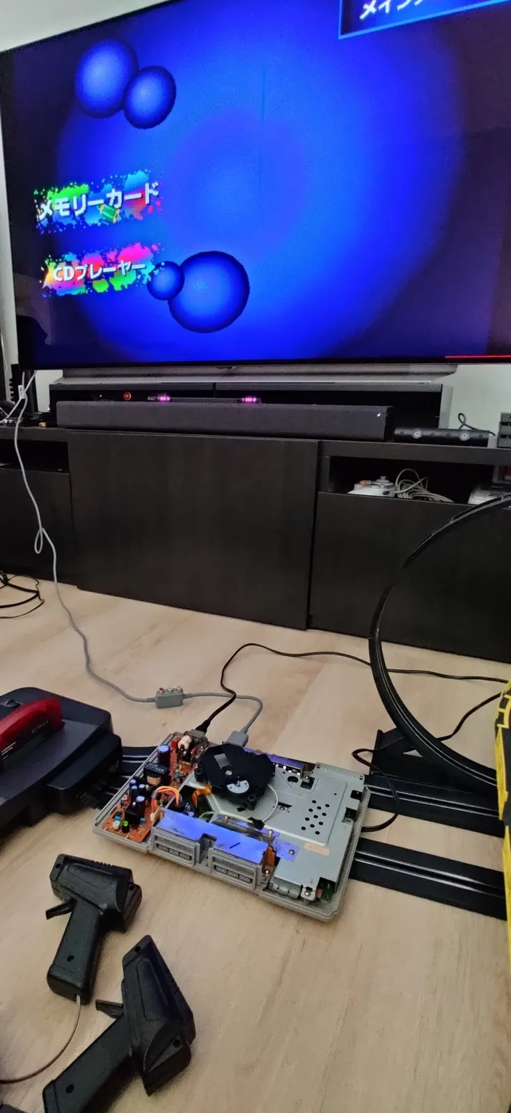 Opened-up NTSC-J PS1 on the floor in front of a TV showing the Japanese PlayStation menu with no game loaded (Memory Card and CD Player options on screen). Nearby are pistol-grip controllers for a slot-car track and a loop of the track, and to the left sits a charcoal Nintendo 64 with a red Krikzz Everdrive 64 X7 flashcart in its slot.