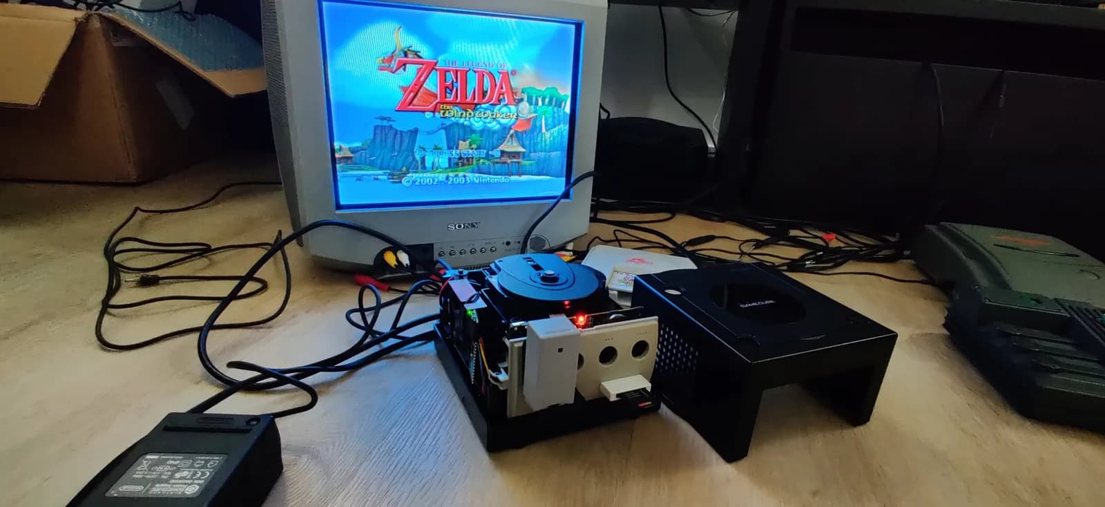 An opened black Nintendo GameCube (top shell removed) powered on, its optical drive still fitted and the green PicoBoot mod board visible in profile on the left by the cooling fan with its status LED lit, and a white WiiKey SD-card adapter (in the front memory card port) holding a red SD card. The removed top half of the shell sits to the right; a Sony CRT behind shows the Legend of Zelda: The Wind Waker title screen.