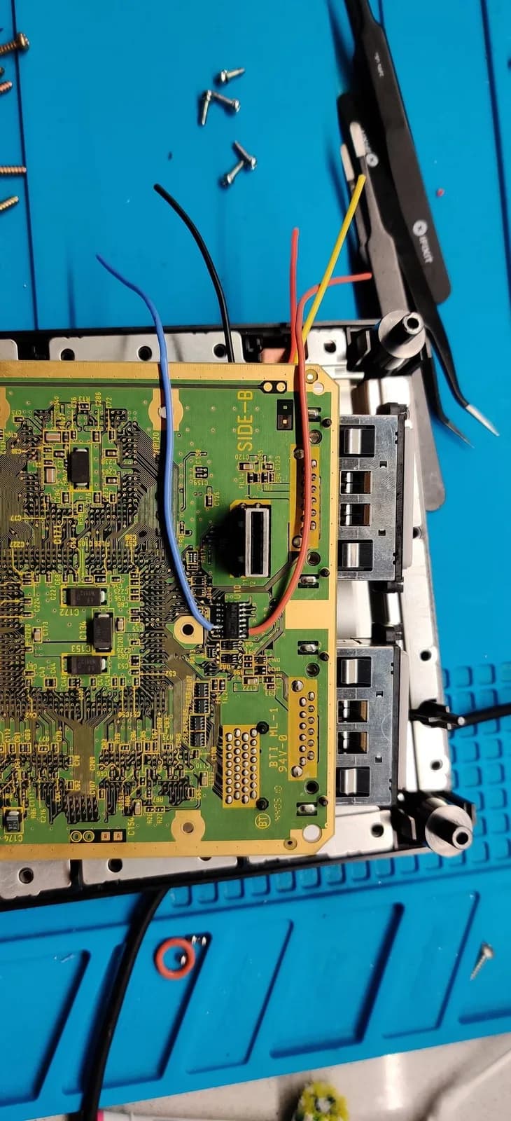 Underside of the GameCube mainboard (SIDE-B silkscreen) during the PicoBoot install, with five mod wires soldered on (blue, black, red, yellow and orange), tweezers, and scattered screws on a blue work mat.
