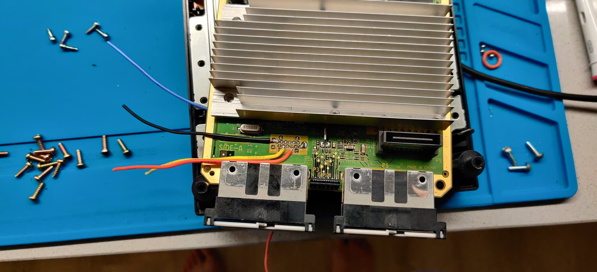 GameCube mainboard undergoing PicoBoot mod, showing green PCB with WiiKey interface, aluminum heatsink, capacitors, and scattered screws on blue workmat.