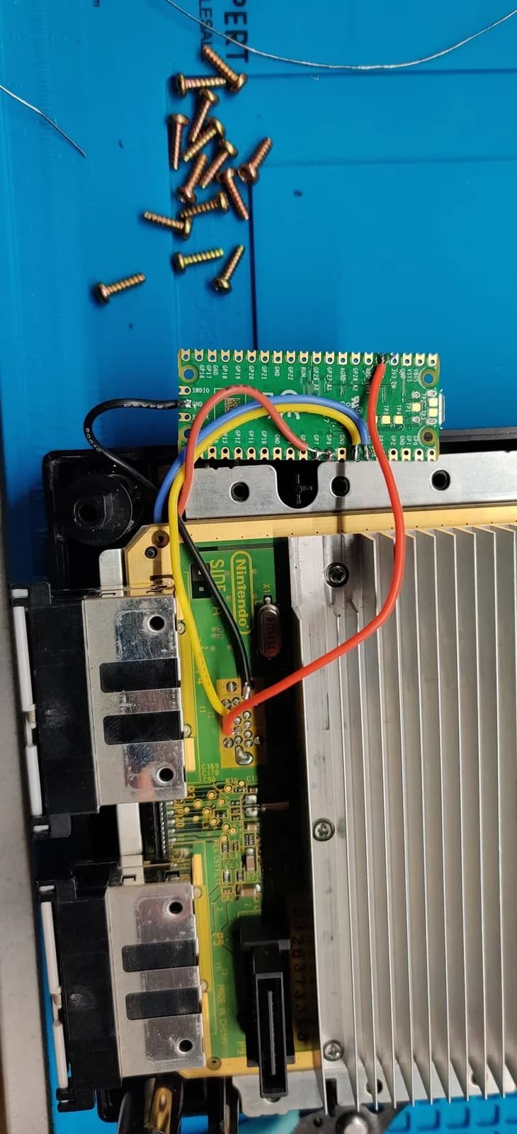 Scattered brass screws above the GameCube mainboard with the green PicoBoot mod board installed, wired in with five wires (blue, black, red, yellow and orange), the aluminium heatsink visible.