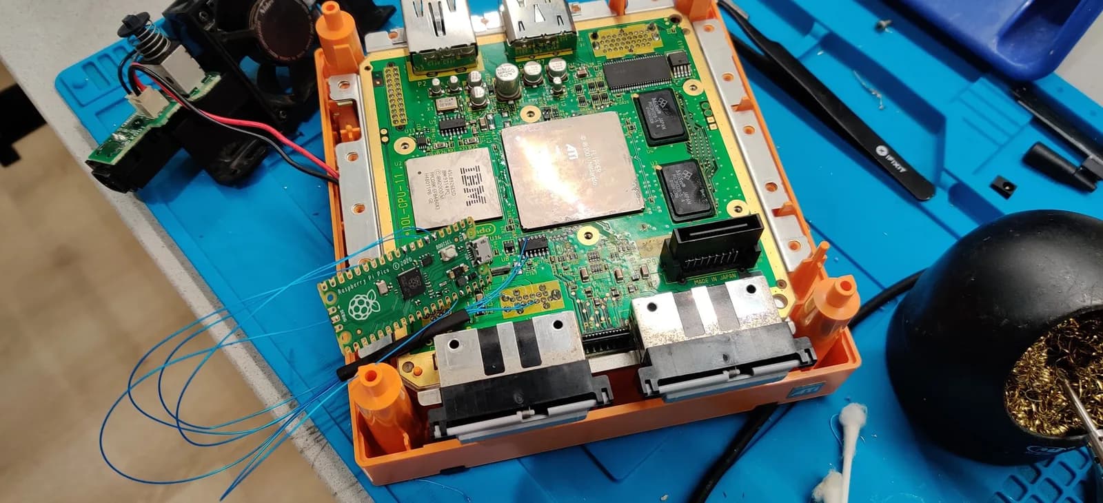 Spice orange GameCube opened on a blue work mat, exposing the green mainboard with a Raspberry Pi Pico (PicoBoot) wired in with blue kynar wire, its install wirework on show.