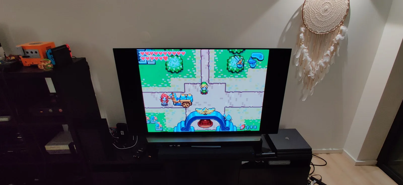 A flat-panel TV showing The Legend of Zelda: The Minish Cap, running on the spice orange GameCube's Game Boy Player via the GBI homebrew (loaded through Swiss, enabled by the PicoBoot), with a silver GameCube, a Nintendo Switch and a PlayStation on the dark media units around it.