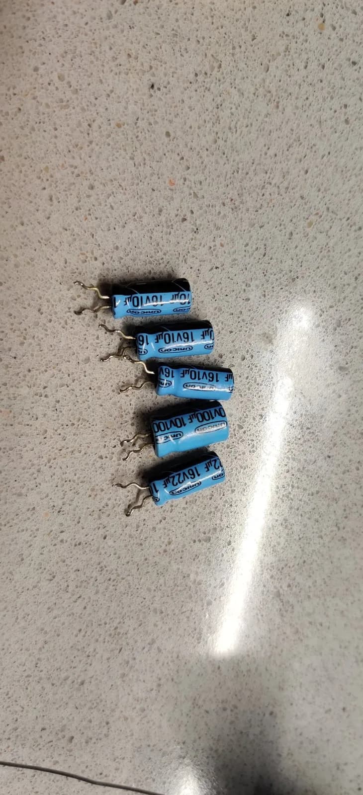 Five old blue electrolytic capacitors removed from the Sega SG-1000 II, legs bent, lined up in a row on concrete.