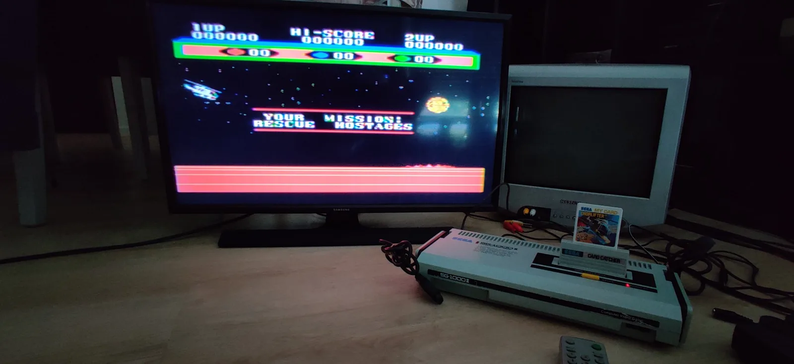 Sega SG-1000 II on a wooden surface running Choplifter, with its 'Your Mission: Rescue Hostages' briefing shown in colour on a Samsung flat-panel TV behind it. The small CRT to the right is powered off because the RF-only console only came up in black and white on it. The game runs from a Sega My Card inserted into a Card Catcher adapter, with a remote in front.