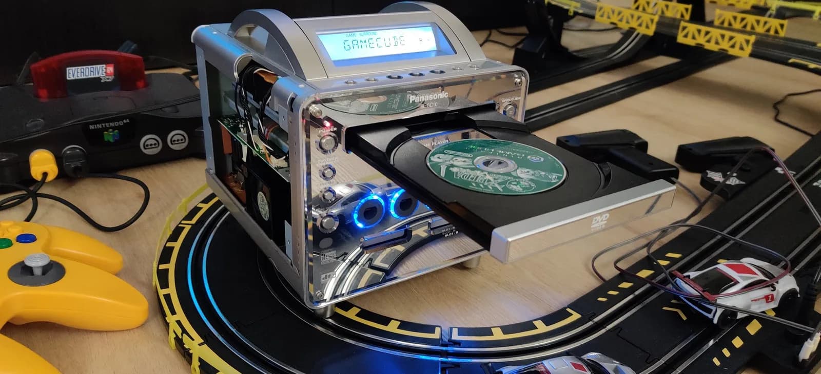 Panasonic Q with open optical drive showing blue LED, disc tray extended with game visible, yellow controller and GameCube console nearby on track racing set.