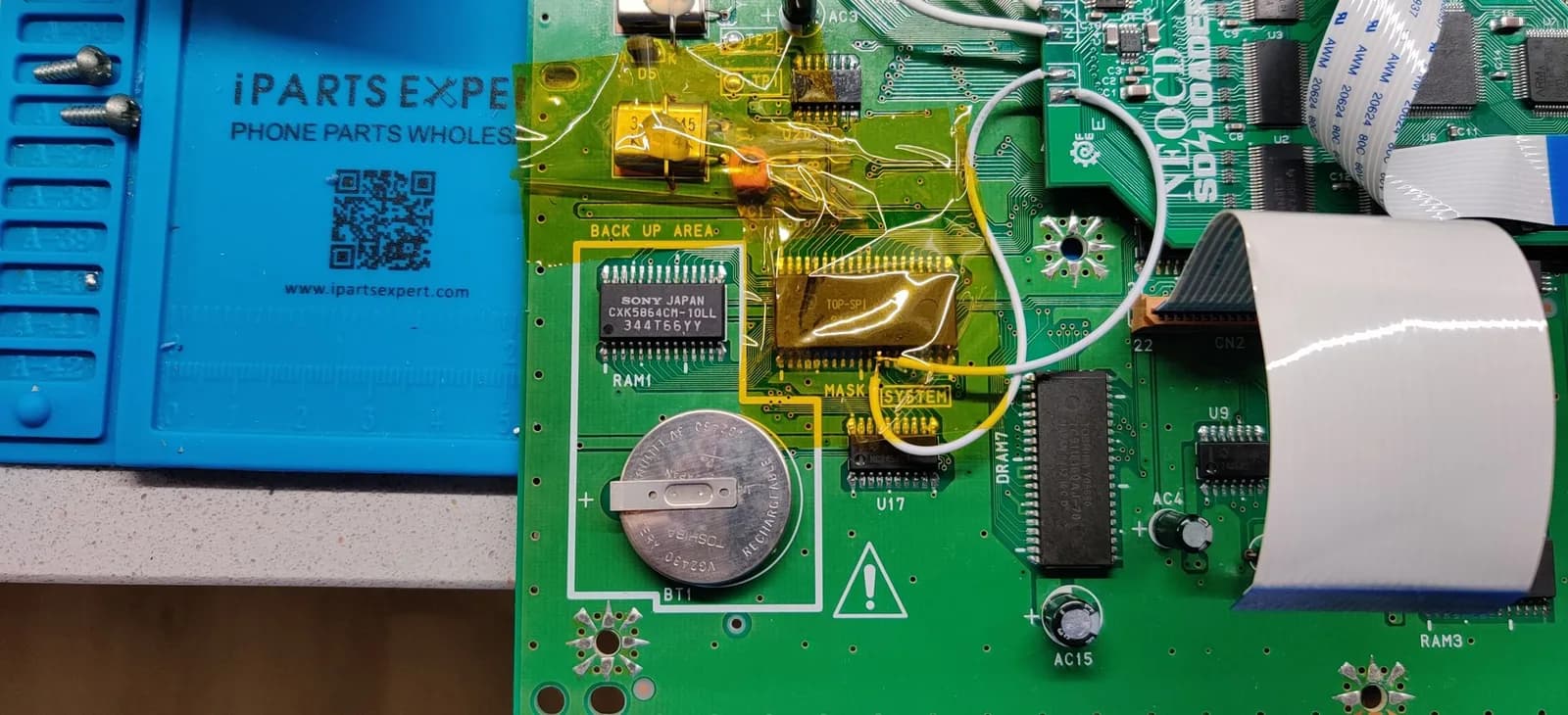 Neo Geo CD mainboard close-up on a blue iParts Expert mat, the save SRAM (SONY JAPAN CXK5864CM-10LL) still in its RAM1 footprint inside the 'BACK UP AREA' silkscreen with the coin-cell battery alongside, the surrounding components masked with yellow kapton tape to shield them from the hot air used to desolder the old SRAM, and the green Furrtek SD Loader board with its ribbon cables at top right.