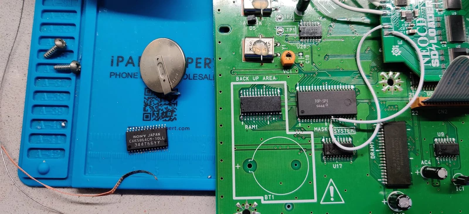 Neo Geo CD mainboard after the save mod: a pin-compatible FRAM chip soldered into the RAM1 footprint in place of the old volatile save SRAM, with the removed SONY JAPAN CXK5864CM-10LL SRAM and the coin-cell battery sitting on the blue mat nearby, the empty battery footprint and 'BACK UP AREA' silkscreen on the green PCB, and the Furrtek SD Loader board at top right. Saves now survive without a battery.