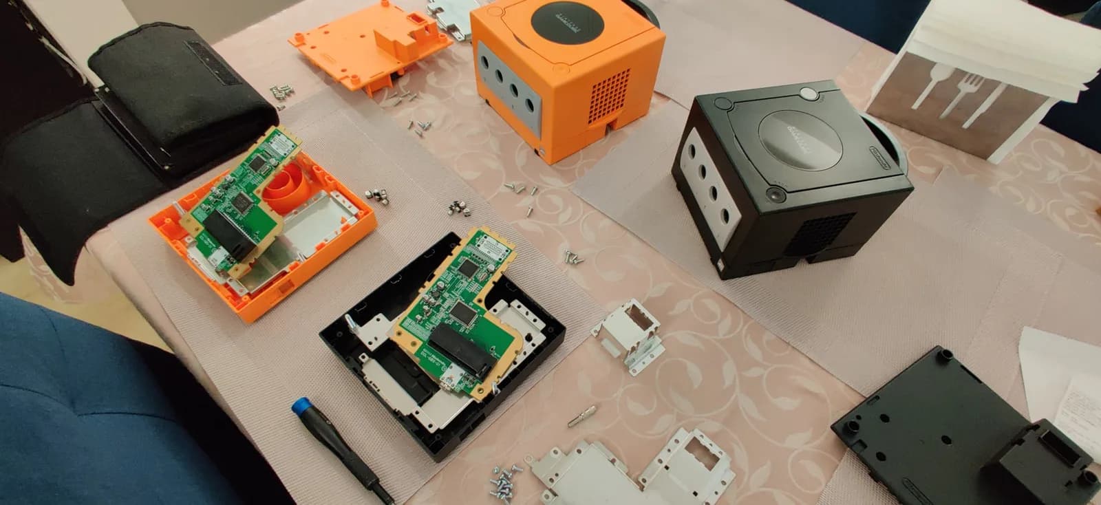 GameCube motherboards and housing components displayed during refurbishment, showing circuit boards with Xeno chip installations and various internal parts laid out for assembly.
