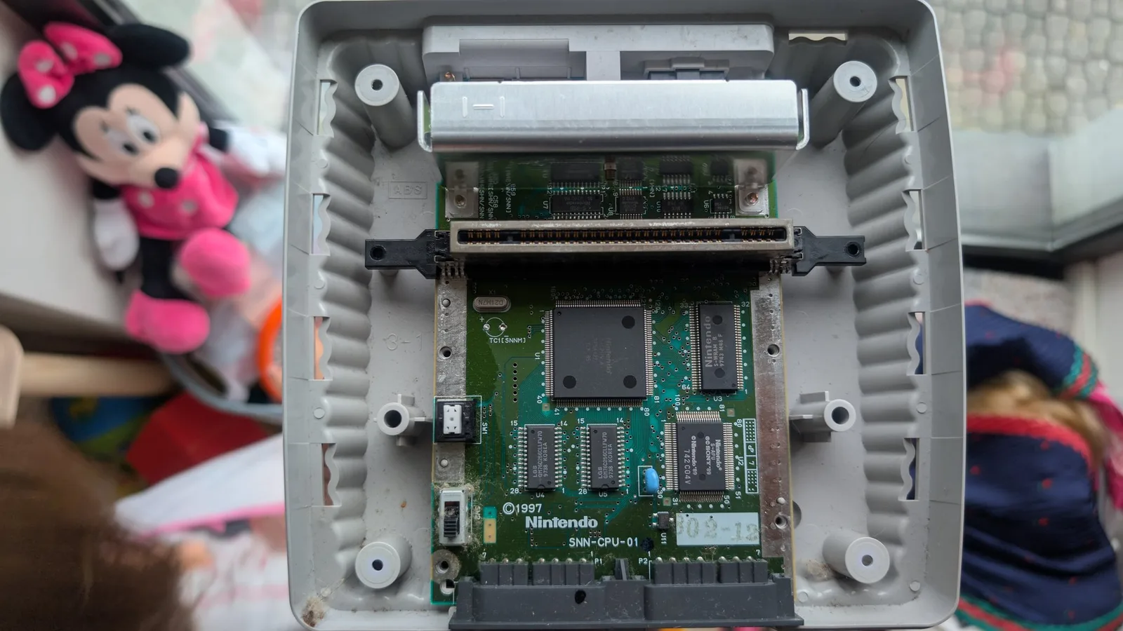 Internal view of Super Famicom Jr. motherboard (SNN-CPU-01) showing recapped circuitry, dust accumulation, and ribbon cable connector during restoration.