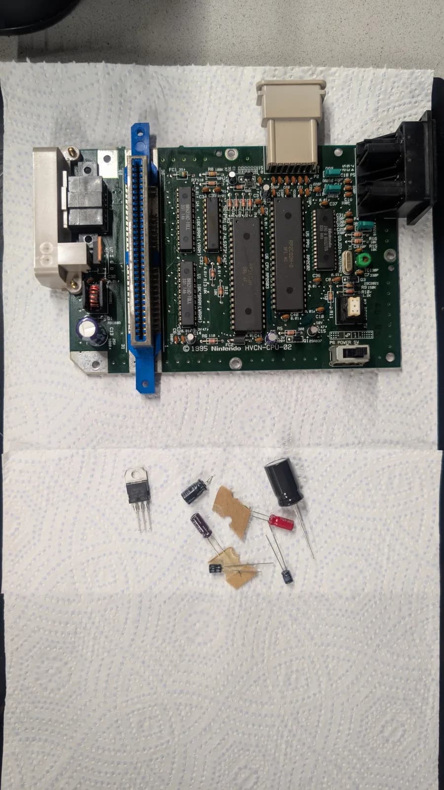 AV Famicom motherboard, component side up, with the new replacement electrolytic capacitors and the new 7805 voltage regulator laid out below it ready to be installed; the cartridge-slot edge connector runs along the board edge.