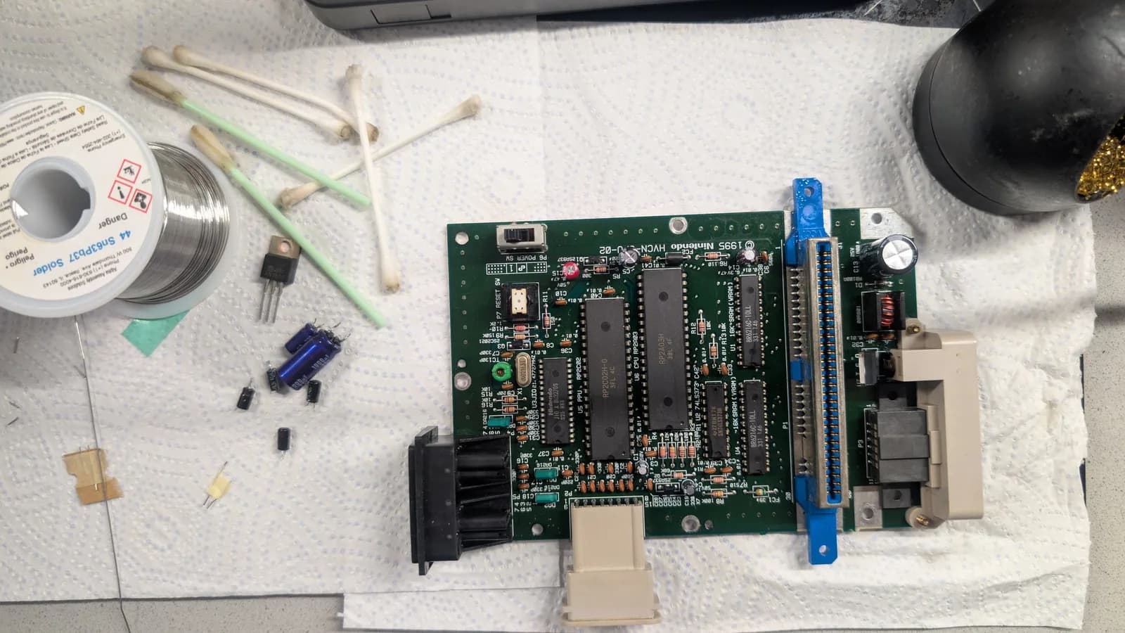 AV Famicom motherboard on a paper-towel work surface, with the old desoldered electrolytic capacitors, a removed 7805 voltage regulator, a spool of Kester Sn63Pb37 solder, used cotton swabs, and a black brass-sponge tip cleaner laid out around it.