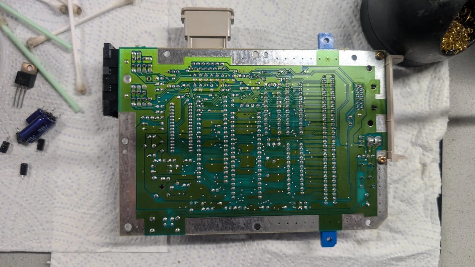 Underside (solder side) of the AV Famicom main board, its copper traces and solder joints exposed; the power jack and the multi-out port were reflowed here. The blue plastic at the edge is the cartridge connector's bracing that screws down into the shell, with a spool of Kester Sn63Pb37 solder, cotton swabs and the brass-sponge tip cleaner nearby.
