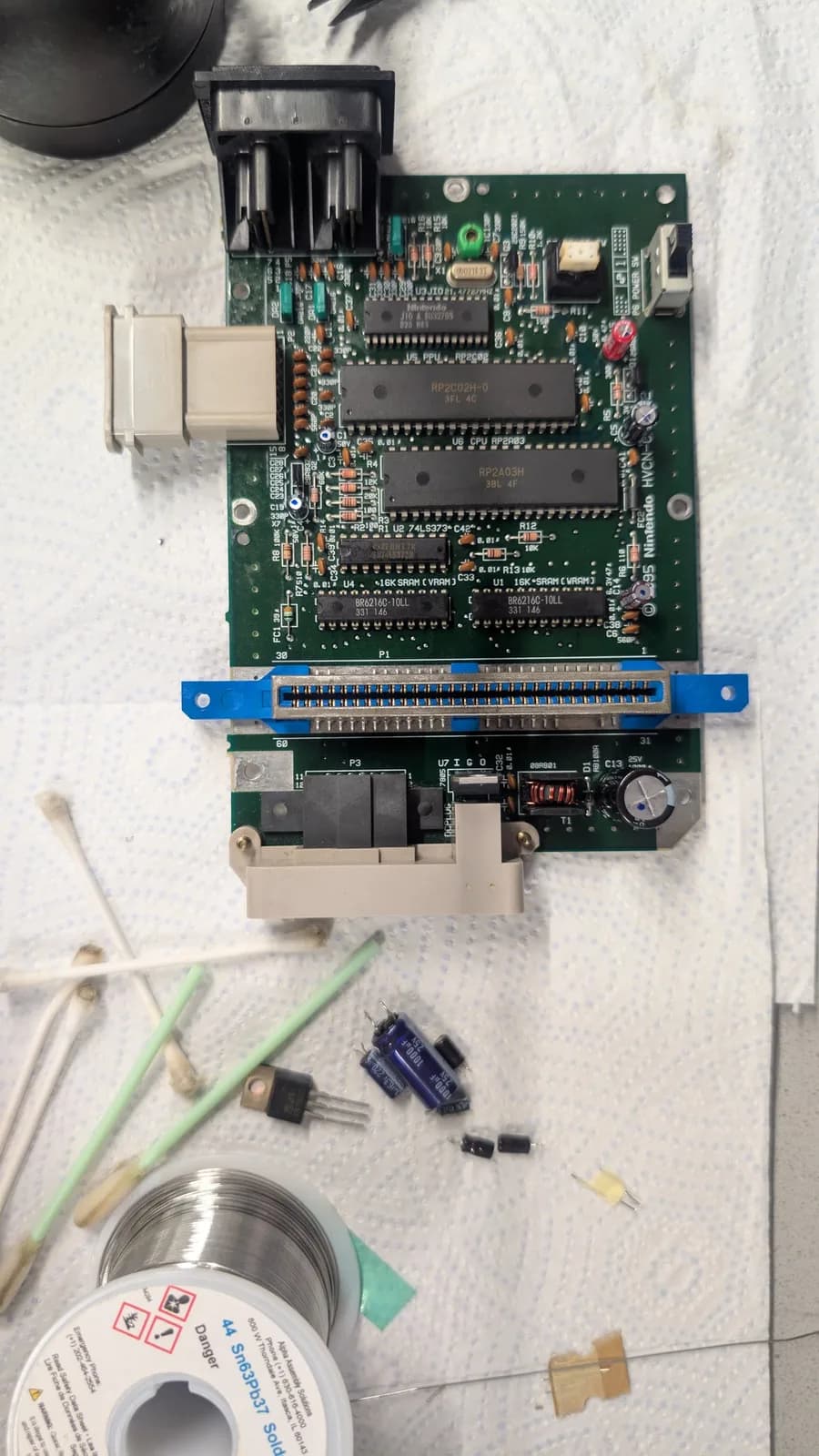 AV Famicom motherboard with the new capacitors installed and each marked with a blue dot, the removed old capacitors and old 7805 voltage regulator laid out below it, alongside a spool of Kester Sn63Pb37 solder, cotton swabs and the brass-sponge tip cleaner.