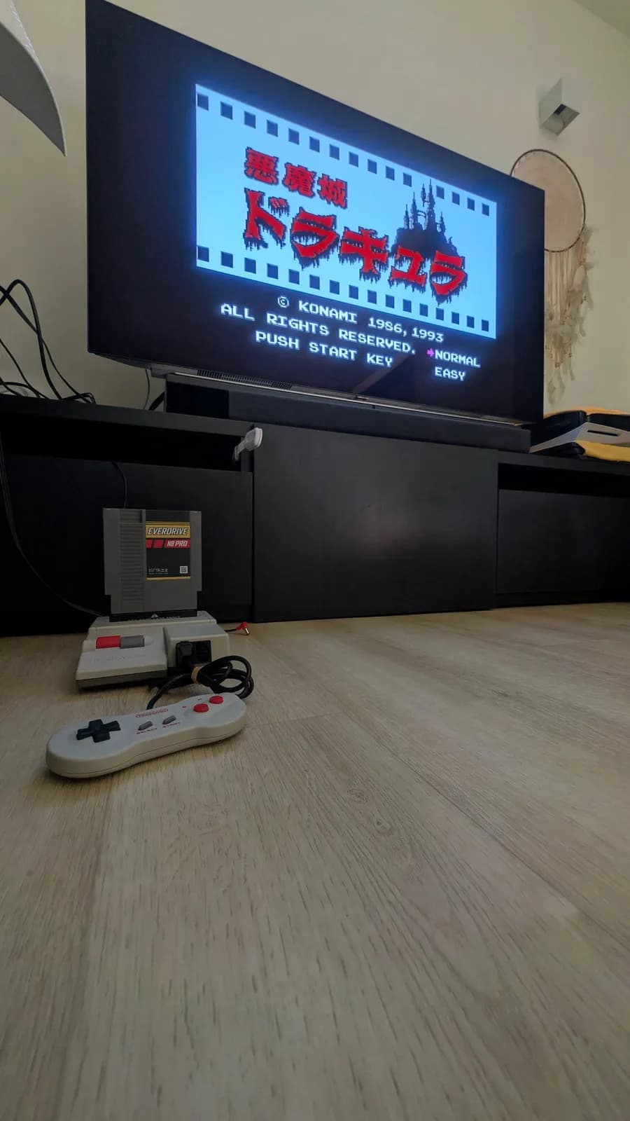 Nintendo AV Famicom (HVC-101) on the floor in front of an LG C1 OLED TV, an EverDrive N8 Pro flashcart inserted and the dogbone controller in front. The TV shows the original Castlevania (Akumajo Dracula) title screen, KONAMI 1986, 1993, PUSH START KEY.
