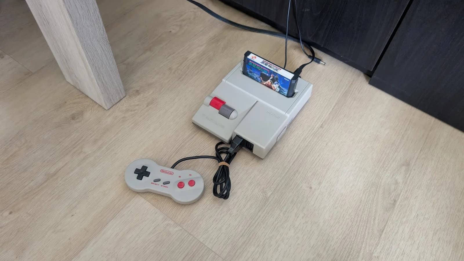 Nintendo AV Famicom (HVC-101) top-loader on a light wood floor, a Konami Castlevania III (Akumajo Densetsu) Famicom cartridge inserted and the dogbone controller with its cord coiled by a rubber band in front, DC power lead connected.