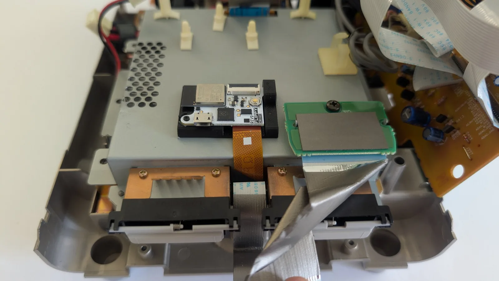 Panasonic Q internals showing FlippyDrive installation with flex cable routed above shielding, green PCB visible, orange copper heatsinks, black drive mechanism.