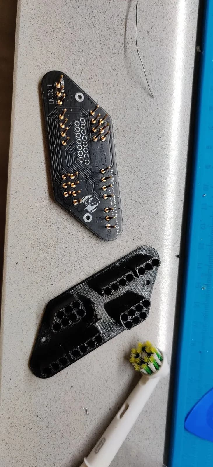 Part of assembling Jeff Chen's TwinDiamond controller adapter: the dark diamond-shaped PCB with gold pins and a 'FRONT' silkscreen, beside its black 3D-printed diamond housing, with an electric toothbrush head for cleaning on a workbench.