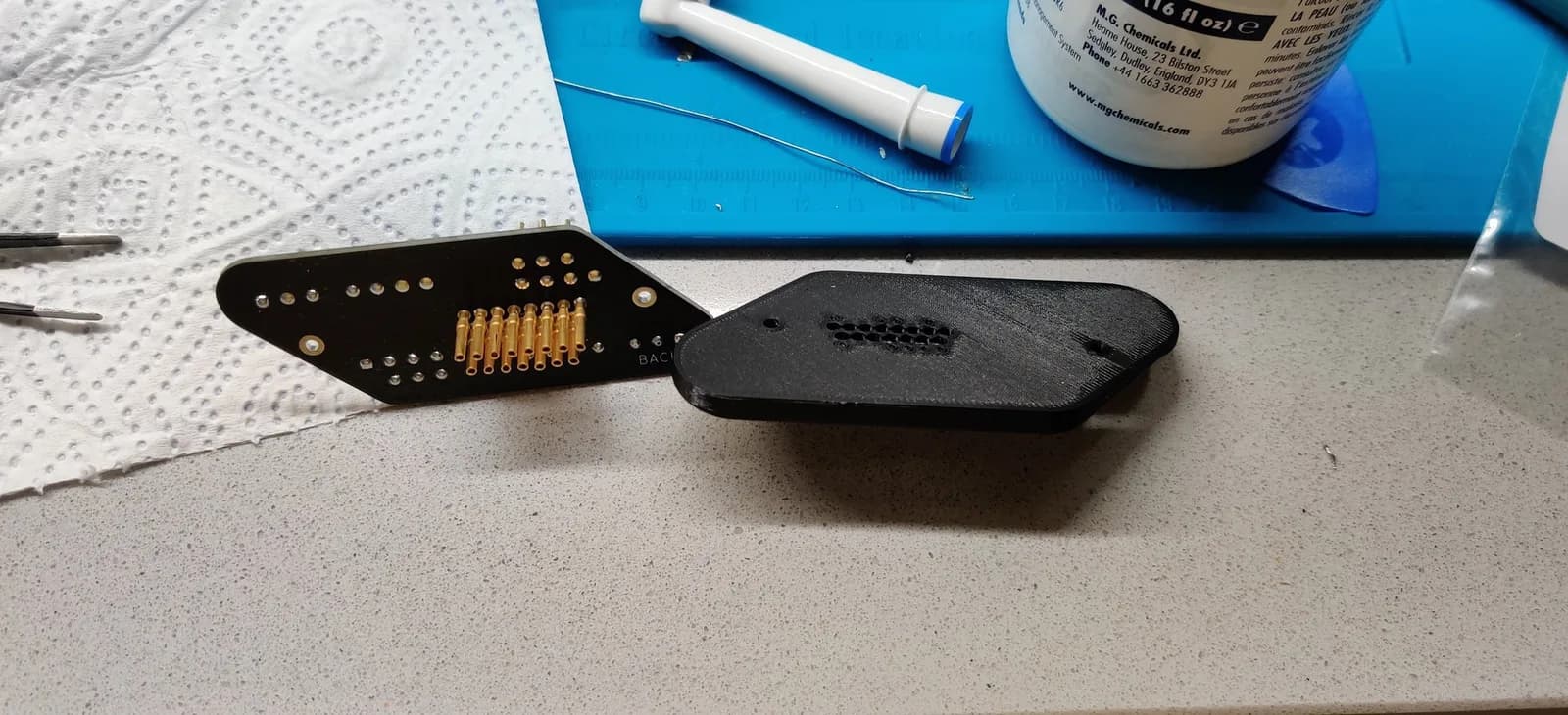 Jeff Chen's TwinDiamond controller adapter, the dark PCB ('BACK' silkscreen) with a row of gold pins, next to its black 3D-printed diamond housing, on a workbench with precision tools and an M.G. Chemicals bottle.