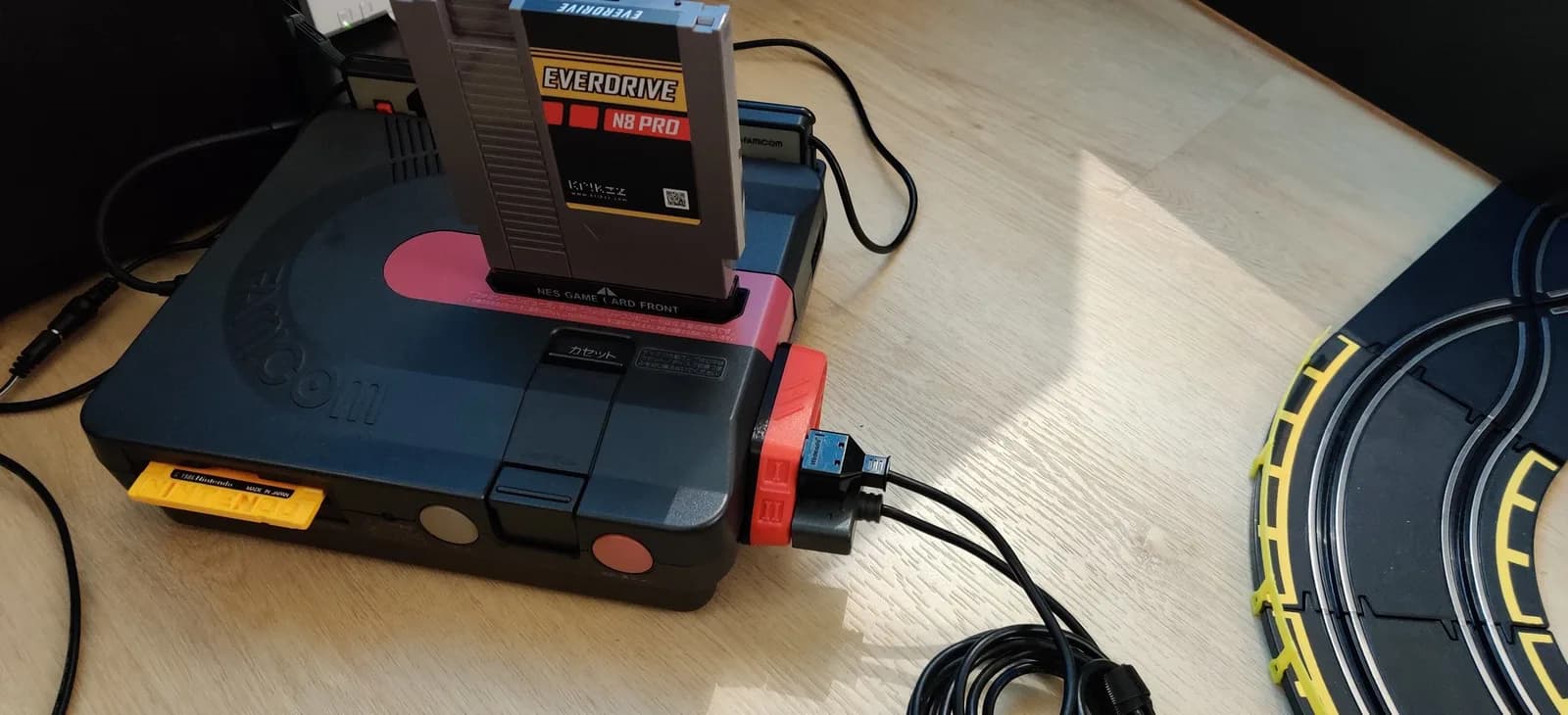 Sharp Twin Famicom (dark/navy AN-500B) on a wood floor with Jeff Chen's red TwinDiamond controller adapter fitted, a Krikzz EverDrive N8 Pro flashcart in the cartridge slot, and a slot-car track alongside.