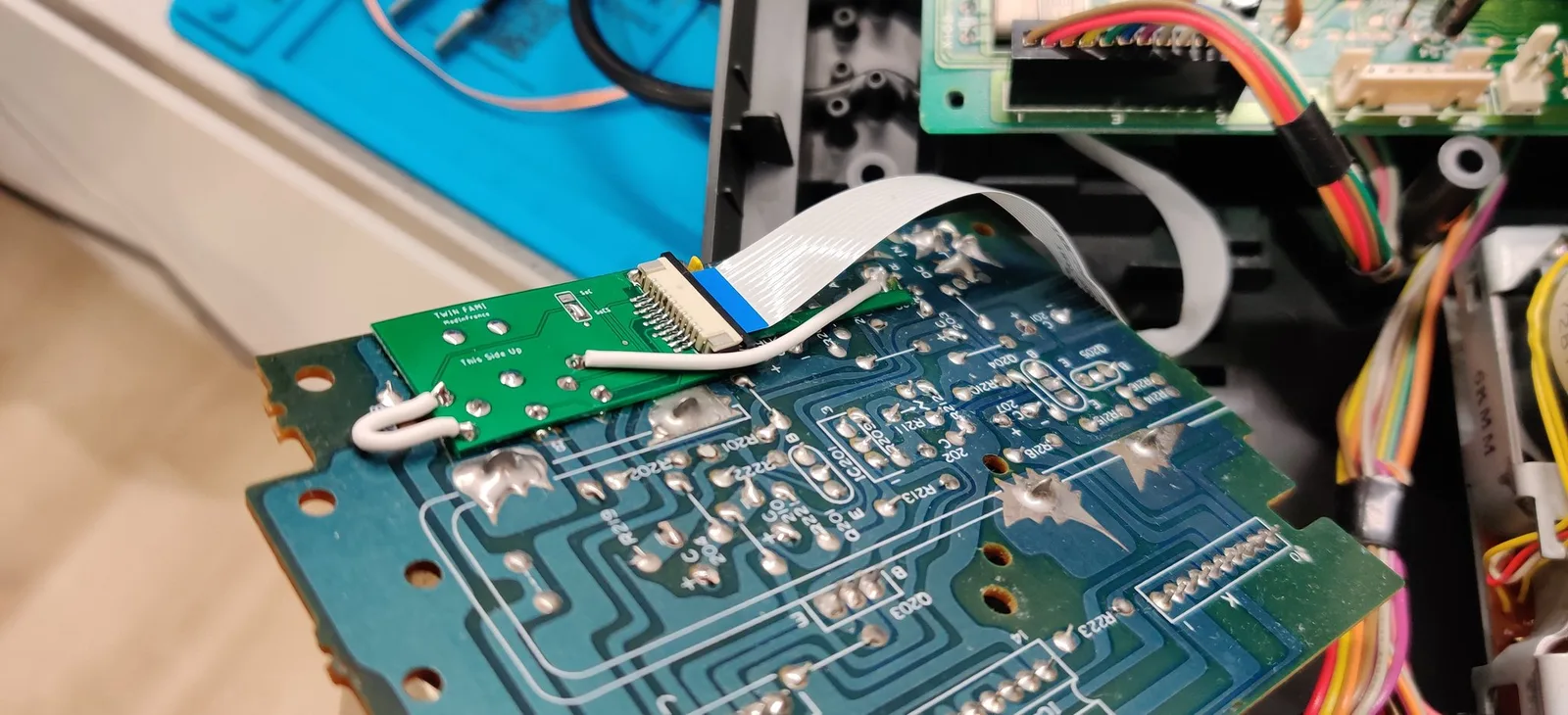 Solder side of the Sharp Twin Famicom board during the NESRGB install. The small green adapter board (silkscreened 'TWIN FAMI / This Side Up') is soldered onto the DIN AV socket that normally carries composite and now outputs RGB, with the ribbon cable from the NESRGB board connected to it, the ModInFrance wireless kit's tidy no-solder-loom way to wire it. A white wire adds a sync-signal fix for a Neo Geo AES RGB cable, a second white wire bodges audio from the RCA jack to the DIN pin so it carries through the RGB cable, and the adapter's SoCS pads are bridged for Sync on Composite Sync. The FDS drive mechanism and wiring looms sit behind.