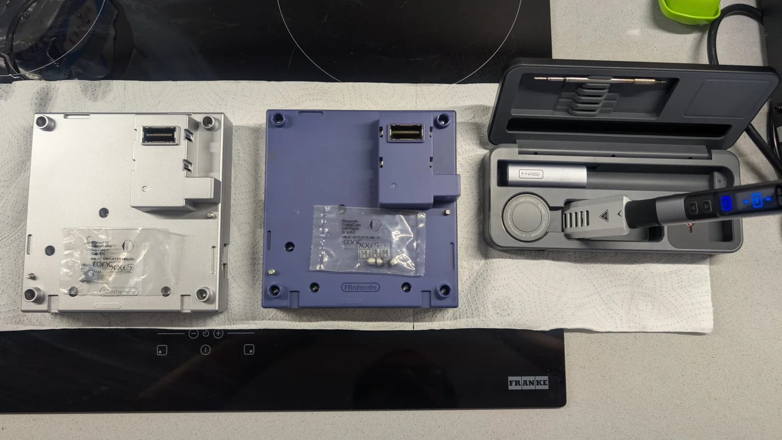 Two Nintendo Game Boy Player (DOL-017) units seen from their top side (the side that docks to the GameCube) on paper towel, a silver one and an indigo one, each with a sealed Console5 GameCube GB Player cap kit bag (NIN-GC-GBPLAYER-SMD-CK) resting on it, and an FNIRSI soldering iron in its case to the right.
