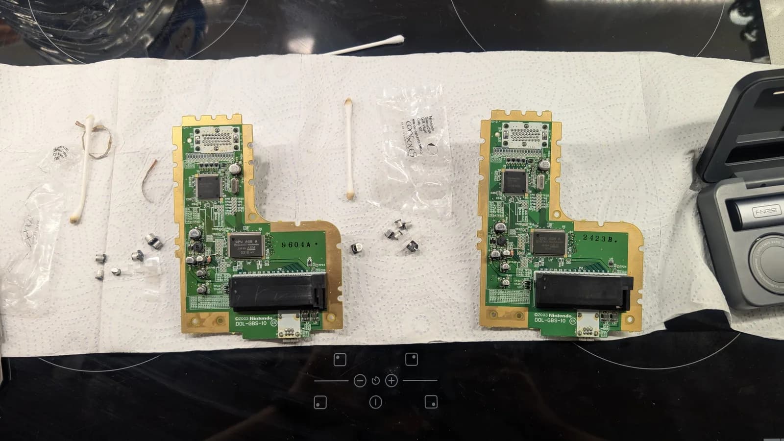 Two Nintendo Game Boy Player boards (silkscreen DOL-GBS-10, CPU AGB A) on paper towel during the recap, the removed old capacitors laid out to the left of each board, with cotton swabs, cut pieces of desoldering braid and a Console5 cap kit bag in shot.