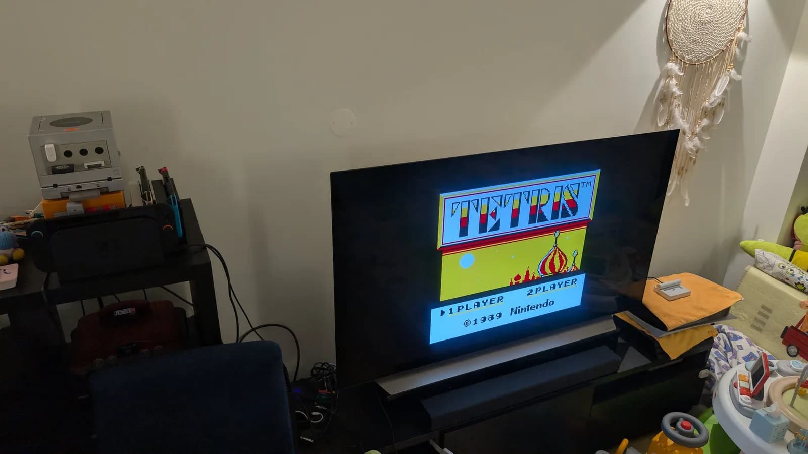 The Game Boy Tetris title screen (1 PLAYER / 2 PLAYER, 1989 Nintendo) on a flat-panel TV through the Game Boy Player. On the stand to the left sit a silver GameCube stacked on a spice orange GameCube, a Nintendo Switch, and a Nintendo 64 with a red EverDrive 64 cartridge.