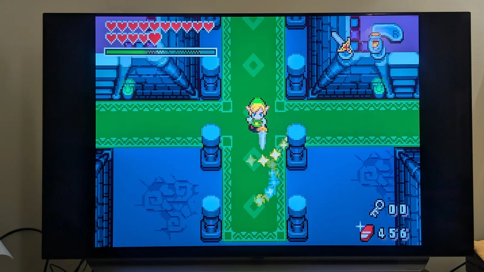 The Legend of Zelda: The Minish Cap running on a flat-panel TV through the Game Boy Player: top-down gameplay of a chibi Link mid spin-attack in a statue-lined dungeon, hearts and a green magic meter up top and 456 rupees.