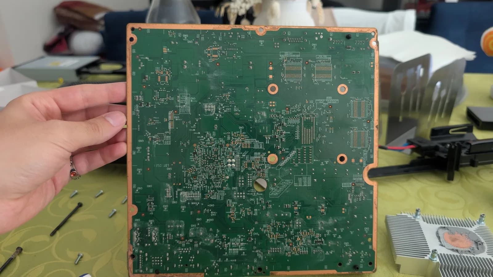 Xbox 360 E motherboard held in hand showing the component-dense solder side with whitish liquid-spill residue and discoloration; SMD components and solder joints visible.