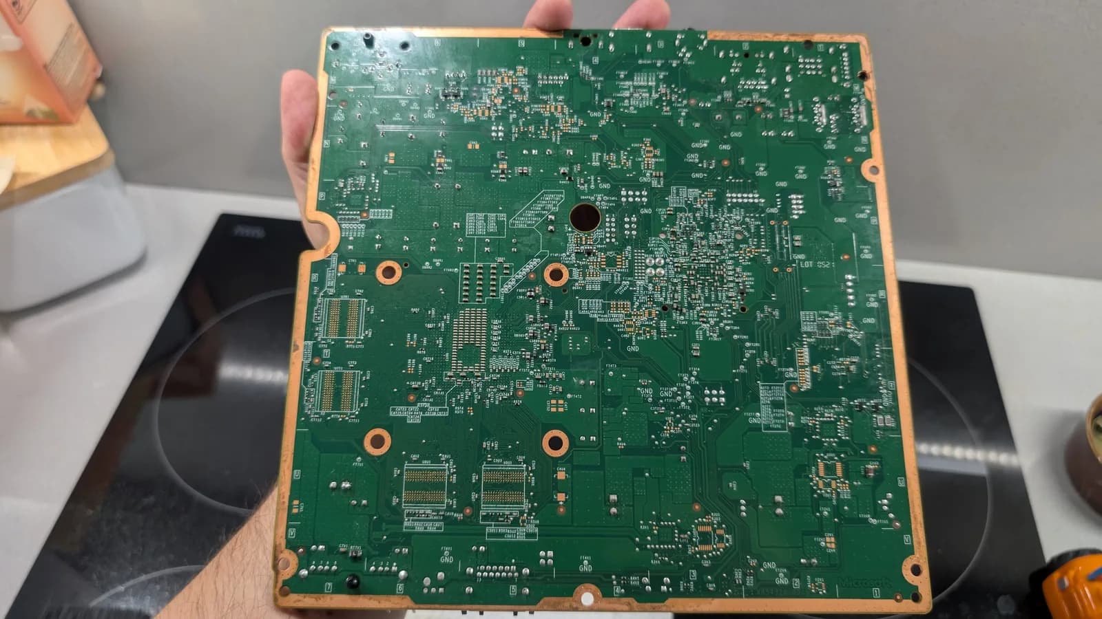 The full Xbox 360 E motherboard (solder side) held in hand over a kitchen hob, some solder joints visibly shiny from the reflow, the board clean after the oxidation was lifted and an isopropyl alcohol wash.