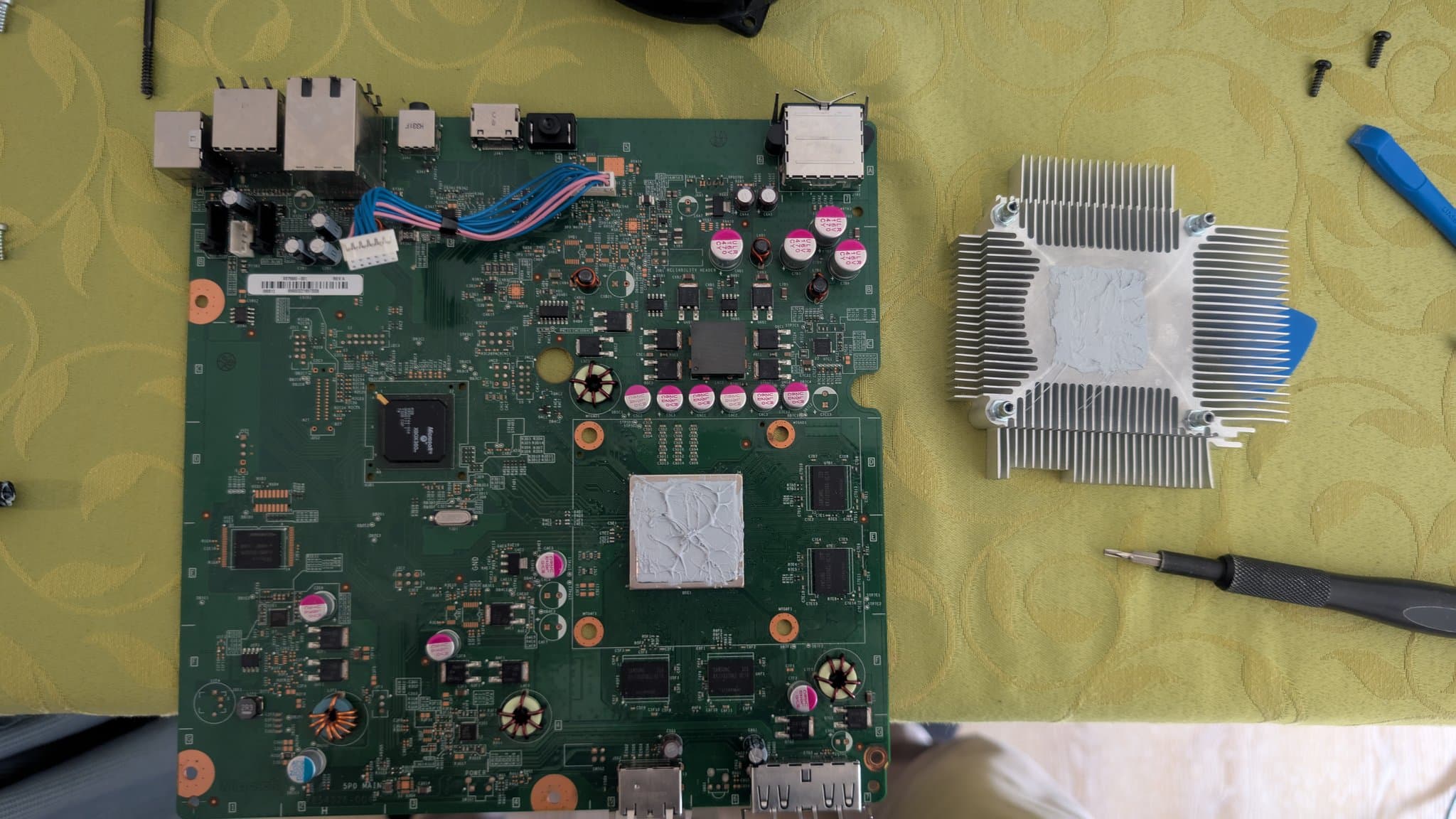 Xbox 360 E mainboard mid-reflow with flux residue, soldering iron nearby, heatsink with fresh thermal paste applied to the right.