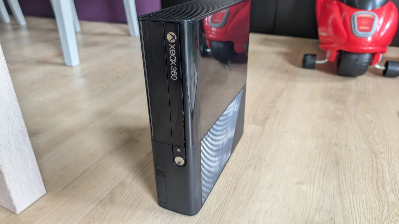 Xbox 360 E console on wooden floor, showing restored black plastic shell with polished finish and reflective surface after cleaning and protective treatment.