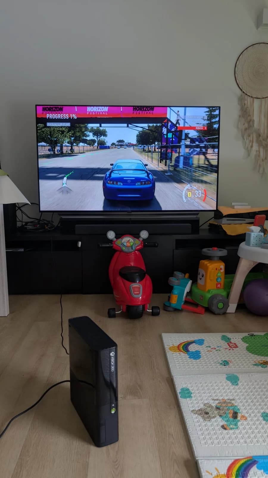 The restored Xbox 360 E standing on the wood floor in front of a black TV stand, the LG C1 OLED TV on the stand showing Forza Horizon, with a red ride-on toy nearby.