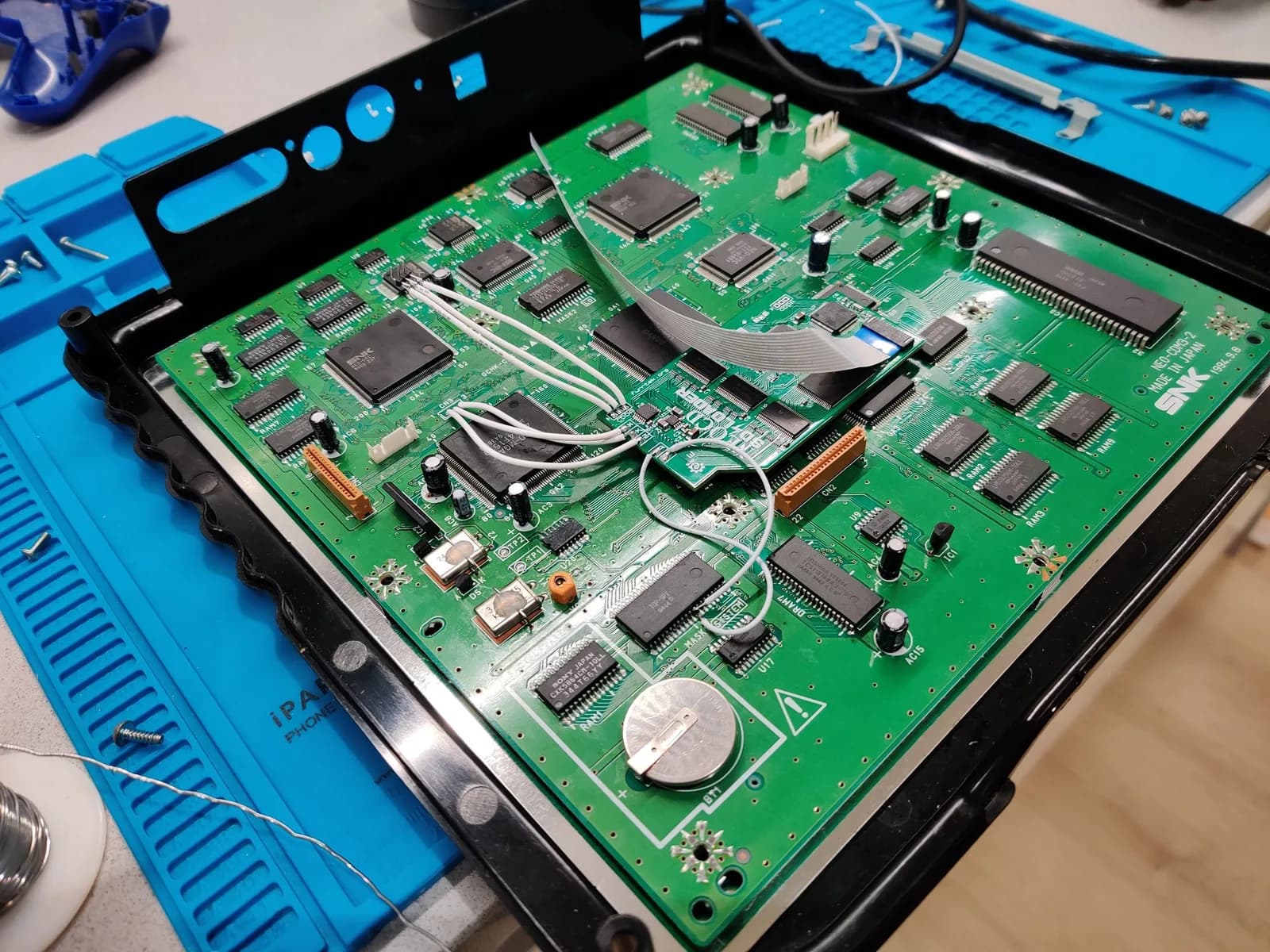 SNK Neo Geo CD mainboard sitting inside its shell on a blue work mat, green PCB with many integrated circuits, a white ribbon cable across the centre, and the white wire work for the Furrtek Neo Geo CD SD Loader mod running across the board, with a coin-cell save battery still fitted near the bottom edge.