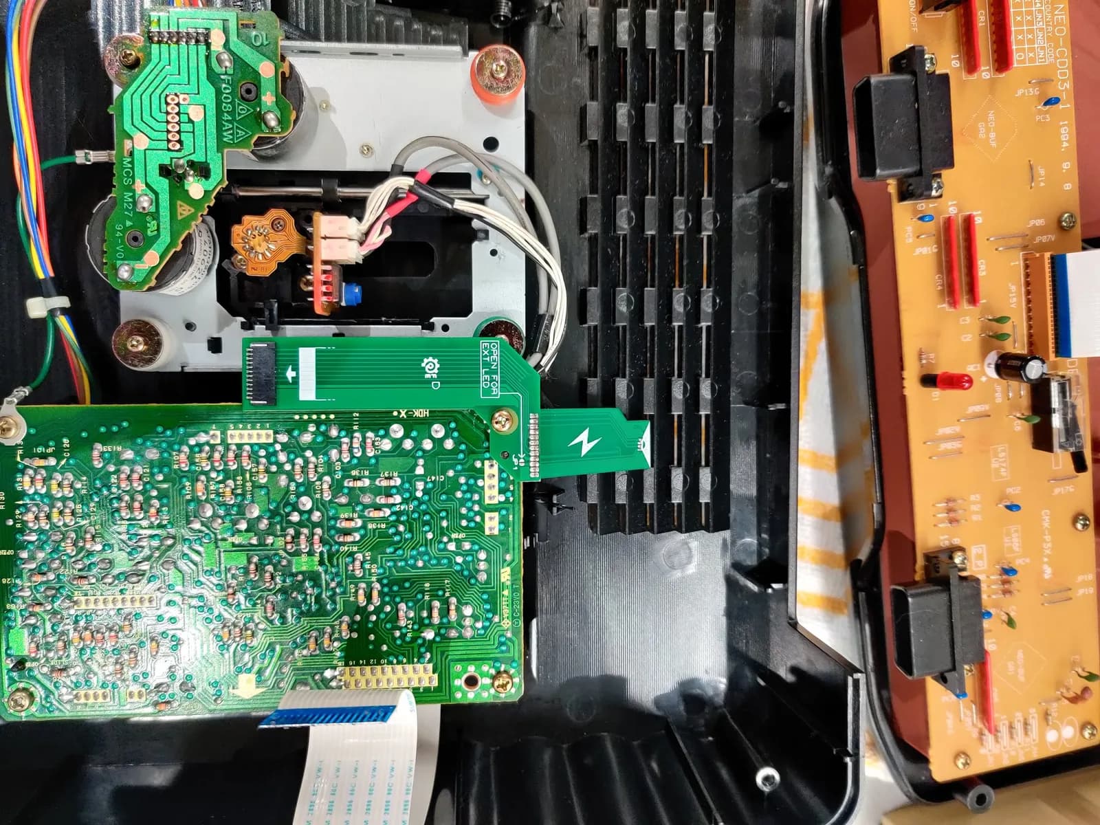 SNK Neo Geo CD opened up showing the drive area: the CD drive mechanism with its laser pickup, the spindle motor soldered to a small green PCB at top left, the micro-SD-card-slot board of the Furrtek SD Loader screwed onto the optical-drive control PCB in the centre (reusing an existing screw post), and the orange controller port board on the right side.