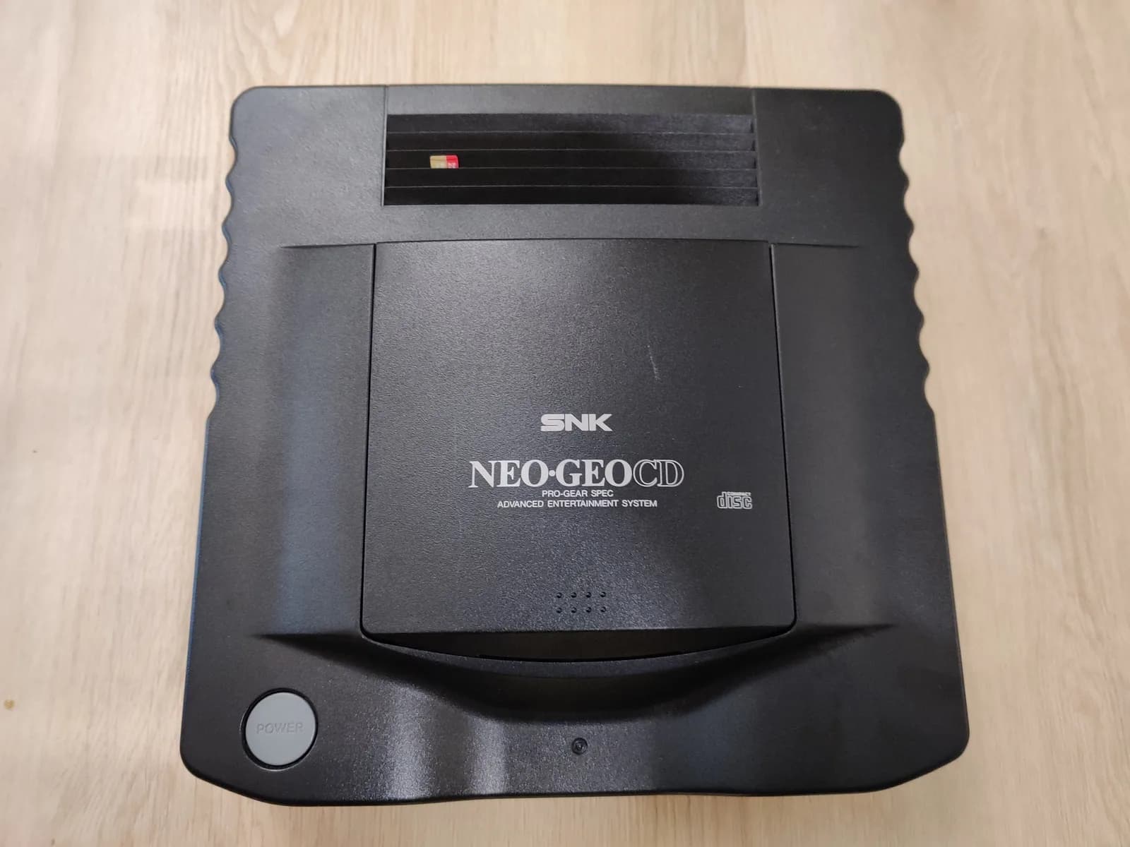 SNK Neo Geo CD console closed and seen from above on a wood surface, matte black shell with the SNK and NEO GEO CD PRO-GEAR SPEC logos on the lid and the round POWER button at the front left, with a sliver of the SD Loader's micro SD card poking out through the ventilation slots in the top of the shell so it can be swapped from outside.