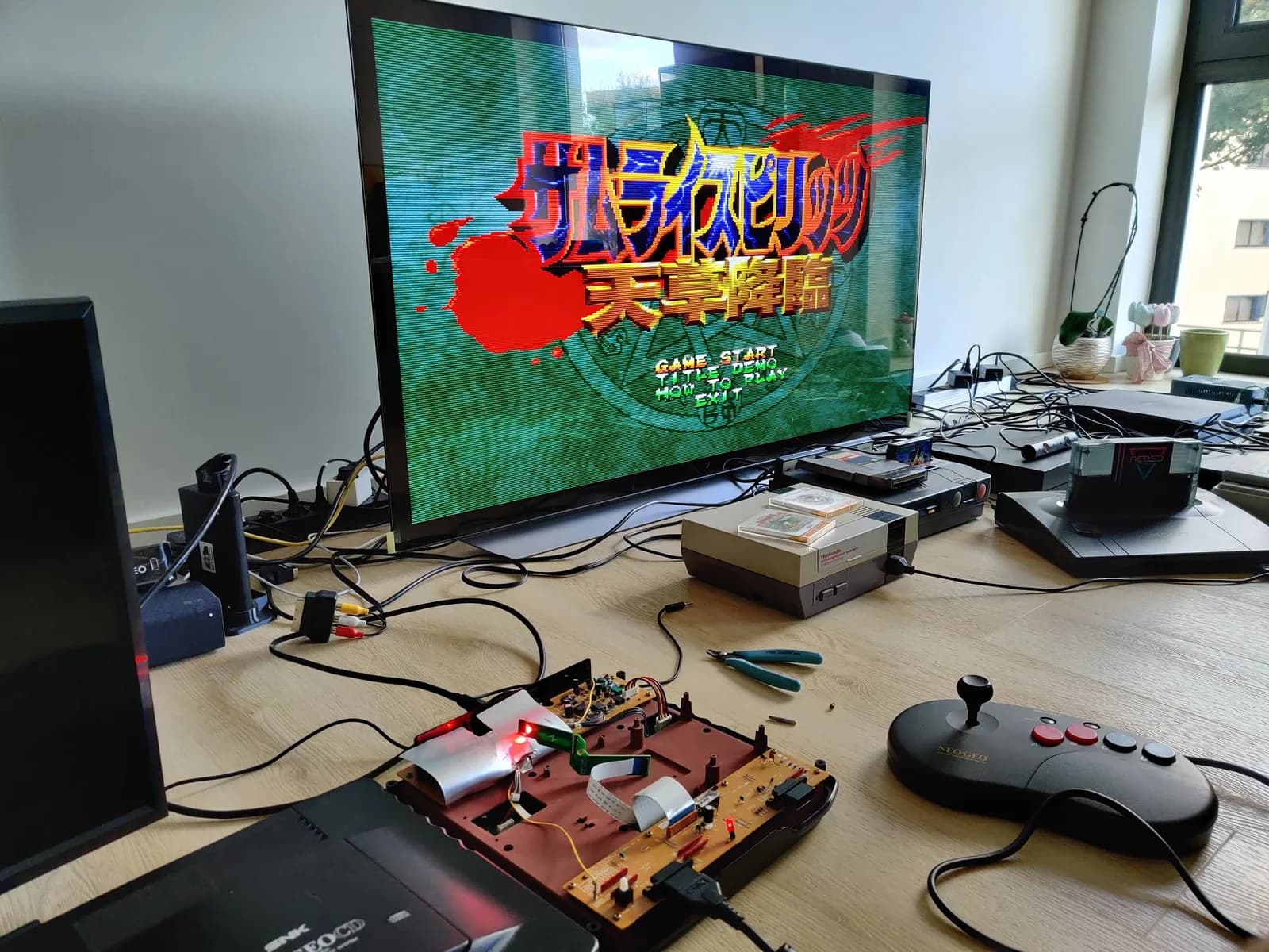Living-room floor testing session: a flat-panel TV showing the Samurai Spirits: Amakusa Kourin (Samurai Shodown IV) title screen, fed from the Neo Geo CD through a RetroTink 5X Pro. The console runs with its top case off, so the CD section is absent, and the exposed mainboard shows two lit red LEDs, one for mainboard power and one on the Furrtek SD Loader showing it working. A black Neo Geo bean-shaped joystick sits in front, with an NES, a Twin Famicom, a Neo Geo AES, an Xbox One X and a PS4 Pro scattered around the floor.