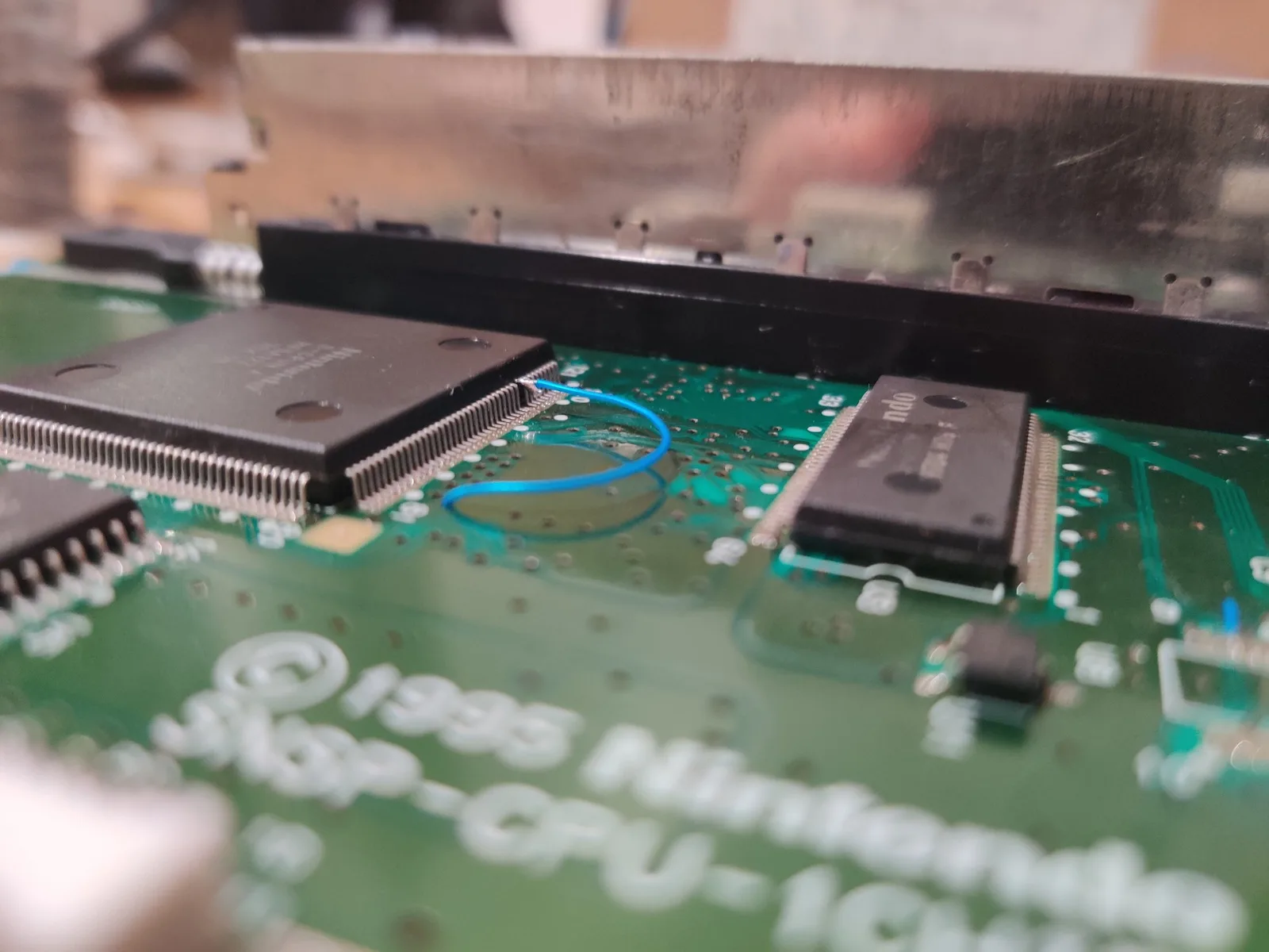 SNES 1-chip motherboard with lifted CPU pins, blue jumper wire, and black heatsink during 50/60Hz mod installation.
