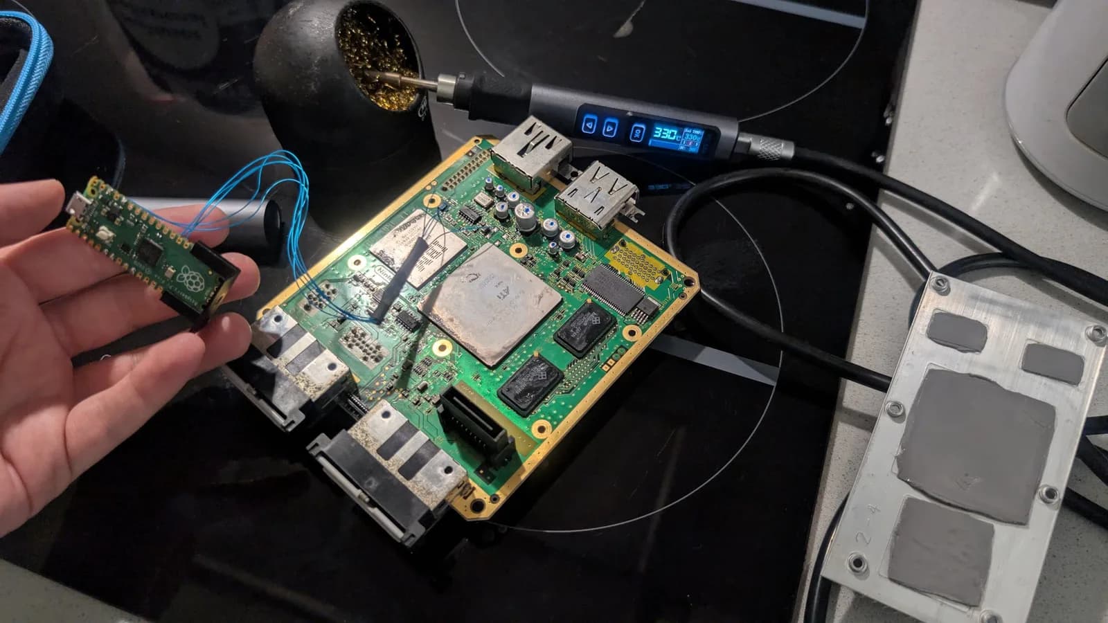 A hand holding the removed Raspberry Pi Pico (PicoBoot, blue wires still attached) over the GameCube mainboard on a black surface, with a portable soldering iron reading 330C and a thermal-pad plate nearby, during the FlippyDrive swap.