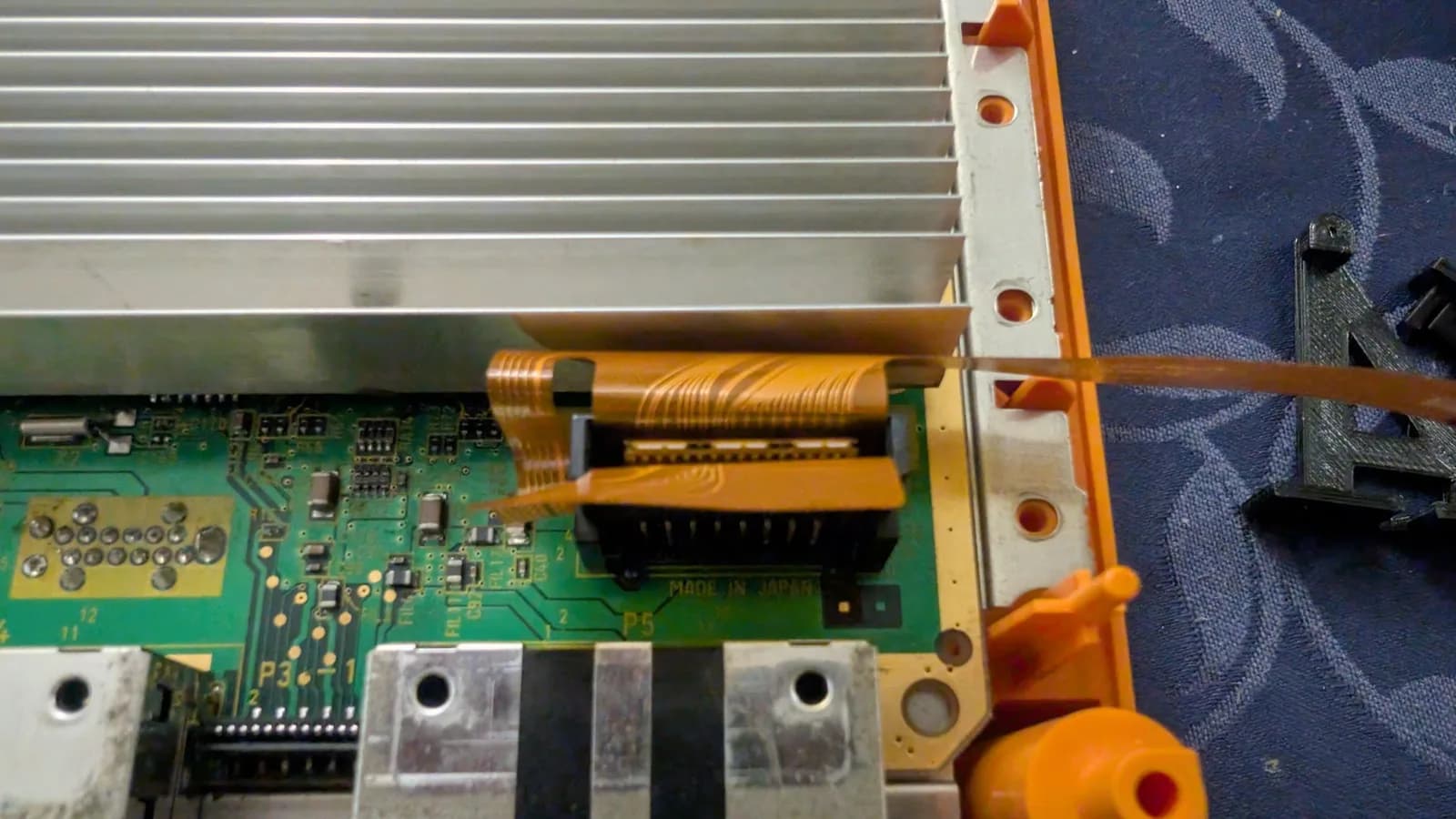 Close-up of the GameCube's optical-drive connector (silkscreen 'P5', 'MADE IN JAPAN') with the FlippyDrive's orange flex ribbon cable properly folded and inserted into it, beside the silver heatsink and the orange chassis, a black 3D-printed bracket on the blue cloth at right.