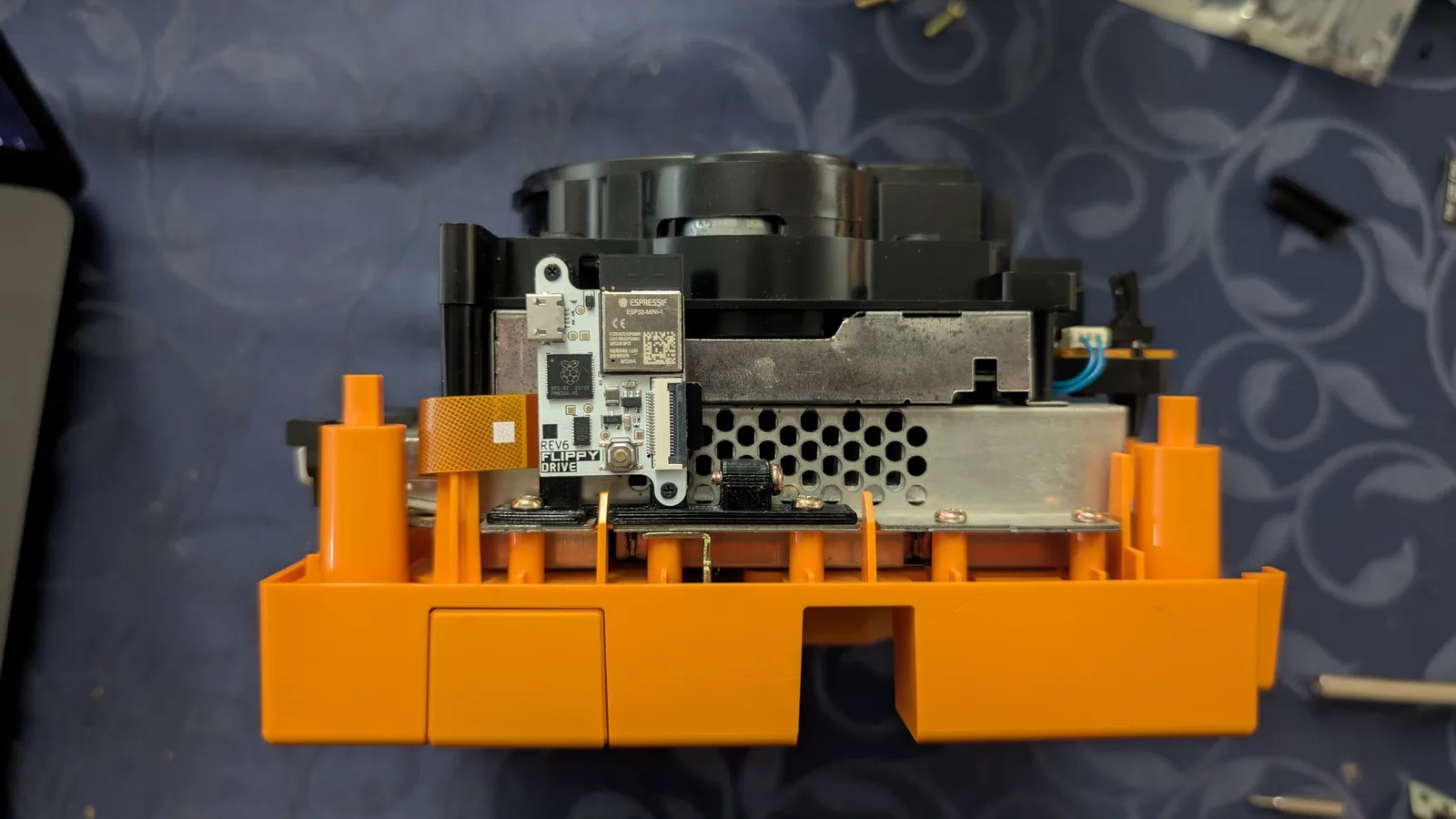 The FlippyDrive board (silkscreen 'FLIPPY DRIVE REV6', an Espressif ESP32-MINI-1 module) installed on its black 3D-printed bracket, screwed down to the GameCube's metal shielding with its orange flex ribbon routed around a screw post; the black drive mechanism and silver shielding behind.