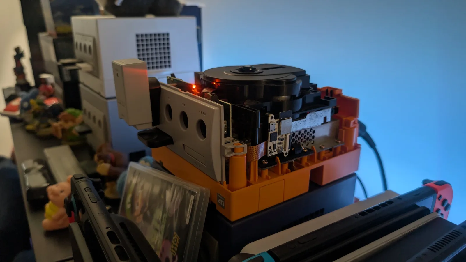 Spice orange GameCube with the top shell off, the optical drive installed in place and pressing down the FlippyDrive's flex ribbon cable; the FlippyDrive sits in a black 3D-printed bracket right beside the drive, its red LED lit. A white console, amiibo figures and a Nintendo Switch are around it.