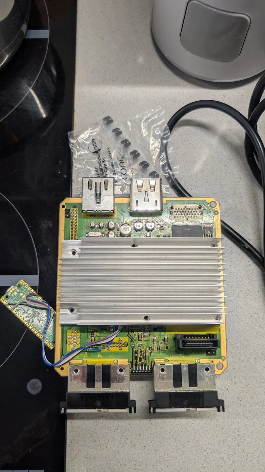 GameCube mainboard about to be recapped, with its large finned heatsink and rear AV-out ports, a Console5 cap kit in a plastic bag above it, and a Raspberry Pi Pico (loaded with PicoBoot) soldered to the board.