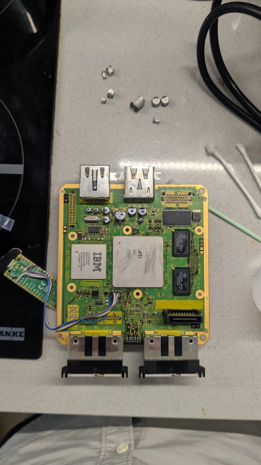 Recapped GameCube mainboard with the IBM CPU, the ATi GPU, RAM and the rear AV-out ports, the new capacitors each marked with a dot and the removed old ones scattered above the board.